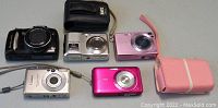 Photo showing five point and shoot digital cameras: Sony Cybershot W310 in black with case, Sony Cybershot W120 in pink with wrist strap, Canon Powershot SX120IS silver with wrist strap, Canon SD300 silver, and Fujifilm AV100 silver.