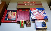 Photo showing all game items together including Monopoly Investor Edition box, Mahjong set, two decks of cards, Skip Bo Deluxe, and Sudoku book.