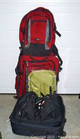 Five pieces of luggage stacked including large red roller case, matching red backpack, unopened olive-green market basket, black carry-on bag, large black shoulder bag