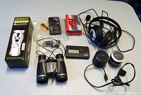 Photo of an emergency wind-up radio/flashlight charger, Panasonic remote control, AAA battery, two unopened Sony cassette tapes, wired headphones, Korg tuner, two mini speakers, and binoculars.