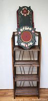 Front view of wooden fold-out shelf showing three shelves with metal decorative support and wall shelf with circular photo space and floral painted decoration on top