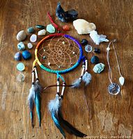 Full lot showing chakra fabric wrapped dreamcatcher with beads and hanging feathers, assorted polished stones, raw crystals, and pendulums arranged on wooden surface.
