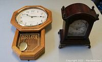 Front view of two clocks: a wooden wall clock with pendulum labeled 'REGULATOR' and a smaller wooden mantle clock with glass front and white dial.