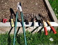 Seven assorted garden tools laid out in a row by a raised garden bed with soil, each tool visible showing handles and working ends.