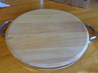 Top view of round wooden cutting board on table, showing light usage on one side and clean surface on the other. Metal handles visible on left and right sides.