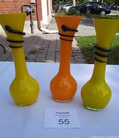 Front view of three colorful glass vases, two yellow and one orange, with black glass spirals wrapped around their necks.