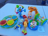 Photo of 7 assorted baby toys placed on a white table outdoors including musical drums, spinning blue monkey, pull dog toy, giraffe rattle, interactive puppy, and additional small toys.