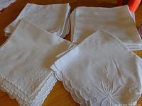 Top view of four folded sets of white cloth napkins showing different embroidery and edge styles on table.