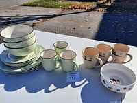 Table display of assorted kitchen dishes including cream-colored bowls and plates with brown rims, cream and light green mugs, and white decorative bowl with colorful circles.