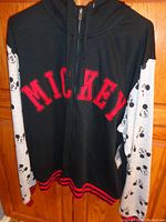 Front view of black hoodie with red embroidered "MICKEY" on the chest and light gray sleeves printed with Mickey Mouse faces hung on a wooden door.
