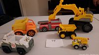 Photo showing all six toy trucks together on a flat surface including the large excavator, yellow dump truck, red fire truck, yellow-orange dump truck, white construction vehicle, and yellow crane truck.