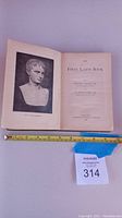 Photo of open vintage book The First Latin Book showing the bust illustration of The Young Augustus and title page with authors and publishing details, measuring tape indicating 7.5 inches width.