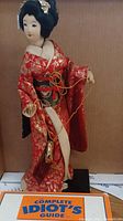 Vintage Geisha doll dressed in red kimono holding purse, mounted on black wood stand with Complete Idiot's Guide to Japanese CD case in front
