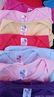 Close-up photo showing seven colorful Strawberry brand baby onesies neatly stacked showing label and colors like pink, yellow, light blue, purple.