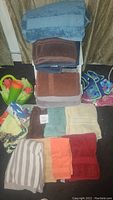 Photo showing stacked bath towels in earth tone colors including blue, brown, beige, orange, red, and striped gray/white, along with visible plastic beach bucket, tumblers, and child floaties with vest.