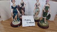 Four hand-painted 1920's lady figurines displayed on a wooden surface, each standing on a round wood base approximately 3 inches wide and 6 inches tall. Figurines lack evident markings.