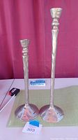 Two pewter tapered candle sticks of different heights, 15.5 and 20 inches, with a hammered texture, placed on a surface against a red curtain background.