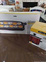 Boxed Bella large electric griddle and boxed Bella 1.5 quart slow cooker crock pot on table.