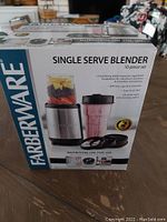 Front view of the Farberware single serve blender box showing the blender with a blending cup filled with fruit, a cup filled with pink smoothie, and blender accessories.