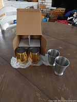 Four metal cups on a wood surface with box and bubble wrap: two gold-toned Mint Julep cups and two silver-toned stemless wine glasses.