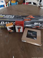 Photo showing the Daytona 2 ton cross beam jack box with branding and lift capacity, along with the digital tachometer, LED light strip box, and toy car packaging all placed on a wooden table.