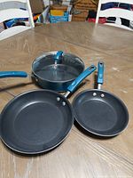Photo showing the set of three cookware pieces on a wooden table: two frying pans and one saucepan with glass lid, all with blue handles.