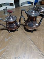 Two silver plated coffee pitchers, one smaller creamer and one larger coffee pot, shown on wooden table