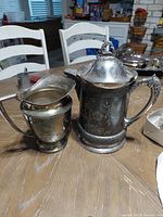 Two silver plated pitchers on a wooden table, one small creamer style and one larger lidded pitcher with floral engraving and ornate handle and lid finial.