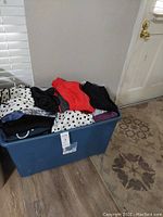 Large blue plastic bin filled with assorted women's pants, shirts, and shorts in various colors including black, white with polka dots, red, and striped patterns.