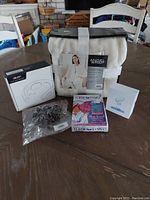 Photo showing all items in the lot together including the Angel Wrap wearable blanket package, neck massager box, Turbie Twist hair towel packaging, scrunchies in a plastic bag, and contact lens cleaner box.