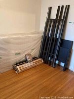 Disassembled bunk bed frame parts leaning against wall next to wrapped mattress. Bed rails, footboards and some slatted supports visible. Hardware package included.