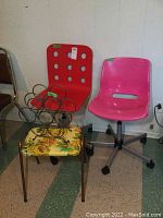 Two rolling office chairs (red with holes, pink plastic) sitting side by side with fabric ottoman, plastic stool and metal wine rack in front