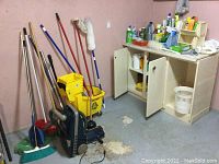 Wide view showing mops, brooms, yellow mop bucket, vacuum, cabinet with cleaning supplies, Tupperware containers, and other cleaning items in basement setting