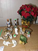 Photo shows all items in the lot including 14 figurines placed on a wood surface and a synthetic poinsettia plant with red petals in gold foil base.