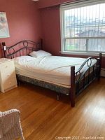 Photo of the full bed showing metal and wood frame, placed in bedroom next to window, with mattress and bedding included