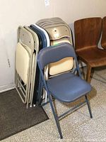 Stack of metal folding chairs in beige, blue, and black colors with padded seats and backs. Chairs stacked leaning against wall with some arranged open in front.