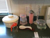 Photo showing a white plastic salad spinner with lid, stacked glass storage containers with pink straw dispenser, white ceramic spoon rest, and paper towel holder on a kitchen counter.