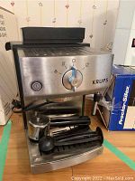 Front angle of Krups coffee machine showing stainless steel front, rotary selector, two portafilters, milk pitcher, and tamper on drip tray.