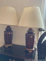 Pair of brown ceramic table lamps with gold accents and beige pleated paper shades placed on a dark tabletop.