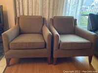 Front view of two brown upholstered MCM armchairs with wooden legs and loose seat and back cushions.