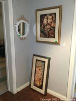 View of corner wall showing one framed painting hanging depicting two human figures, one standing, in warm tones, a circular floral motif mirror mounted on wall, and a second framed elongated painting leaning against wall.