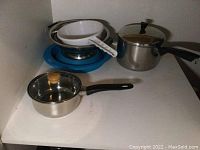 Three pots including small saucepan with lid, two strainers stacked together, and two blue silicone pot holders on white shelf