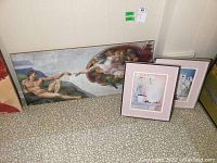 Photo showing three prints on a floor against a wall: a large horizontal Creation of Adam print and two smaller floral prints.