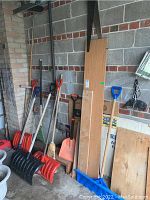 Photo showing six snow shovels and some brooms lined up against a cinder block wall in a garage, including various colors and handle types.