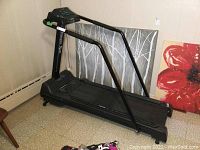 Side view of the black Vitamaster Bio Master treadmill showing metal frame, walking belt, and control panel. Positioned indoors with artwork behind.