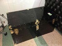 Full front view of black metal camp trunk showing brass hardware and lock.