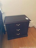 Single wooden side table with three drawers and gold-tone handles shown beside a bed.
