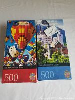 Photo showing two boxed puzzles side by side: Hot Air Balloon Race with red box, Neuschwanstein Castle with blue box.
