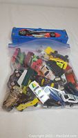Bag of small cars with small blue plastic car-shaped carry case placed on a flat surface, displaying a variety of toy vehicles inside a clear bag.