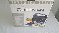Closed box of Chefman Sandwich Grill RJ01 with product image and features listed including 5-minute cooking time, 2 sandwich portions, non-stick coating, and 3-year warranty.